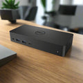 A certified refurbished Dell D6000 USB 3.0 HDMI Display Ports Docking Station by Dell rests on a wooden desk with visible front ports, office furniture and potted plants blurred behind—ideal for versatile multi-monitor setups. Adapter included.